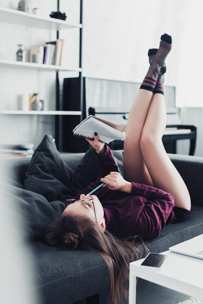 attractive girl lying on sofa and holding notebook and pencil in living room - Foto, immagini