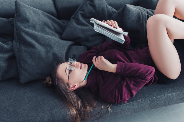 attractive girl lying on sofa and holding notebook and pencil in living room - Photo, Image