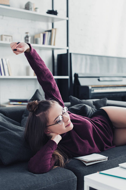 attractive girl lying on sofa, smiling and raising hand in living room - Fotografie, Obrázek