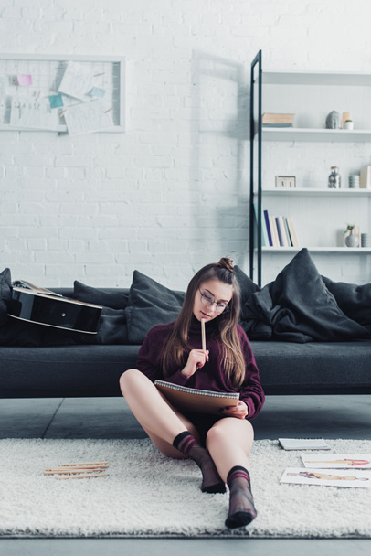 красивый дизайнер сидит на ковре рядом с диваном и держит карандаш и ноутбук дома
 - Фото, изображение