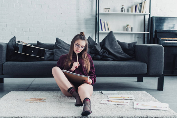 привлекательный дизайнер сидит на ковре рядом с диваном и держит карандаш и ноутбук в гостиной
 - Фото, изображение