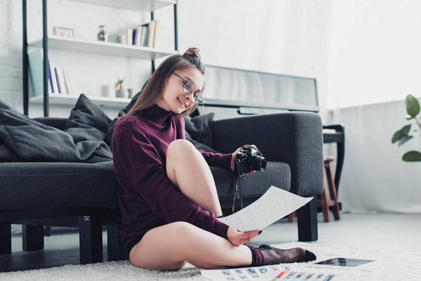 attractive photographer sitting on carpet near sofa, holding film camera and looking at photos in living room - Zdjęcie, obraz