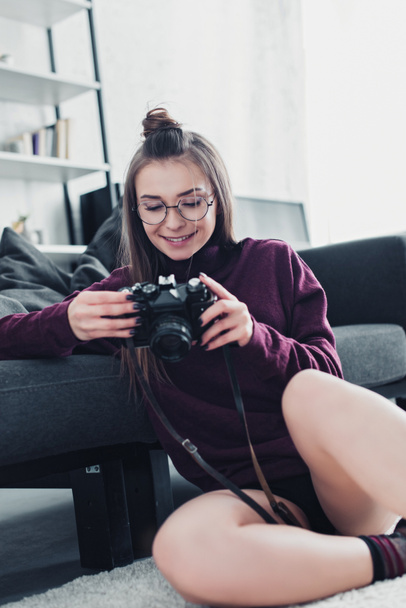 attractive photographer smiling, sitting on floor near sofa and holding film camera in living room - Photo, Image