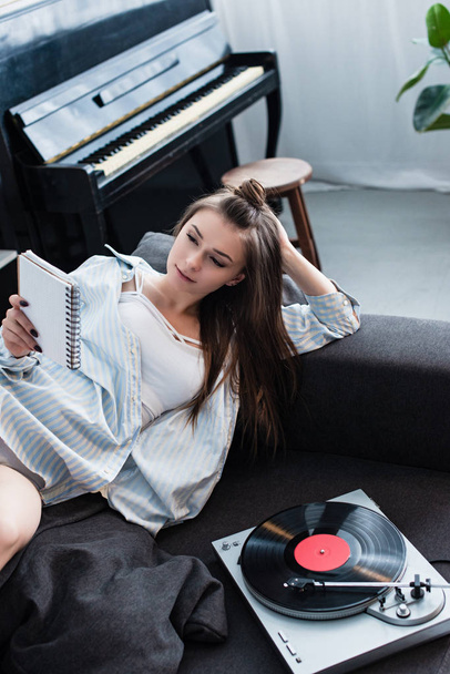 привлекательная девушка лежит на диване возле винтажного проигрывателя и читает тетрадь дома
 - Фото, изображение