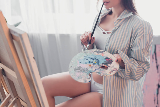 oříznutý pohled umělce drží paleta, kousání štětec a sedí v přední části stojanu v obývacím pokoji - Fotografie, Obrázek