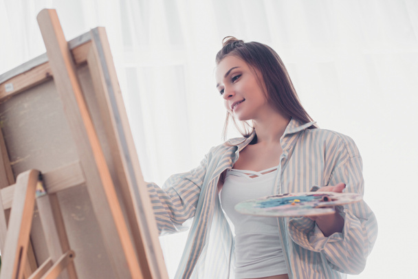 attractive artist holding palette and looking at easel in living room - Foto, Imagem