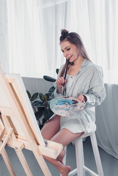 attractive artist sitting on chair, holding palette, biting paintbrush and looking at easel in living room - Фото, зображення
