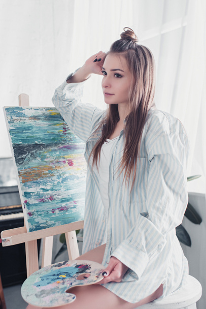 thoughtful artist sitting on chair in front of easel and holding palette at home - Photo, Image