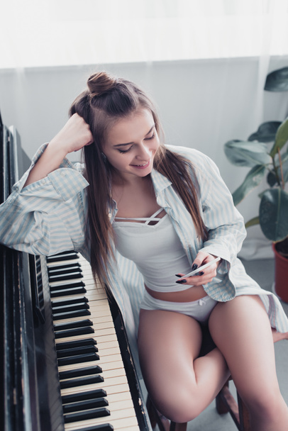 hymyilevä tyttö paita ja alusvaatteet istuu pianon ääressä ja käyttää älypuhelinta kotona
 - Valokuva, kuva