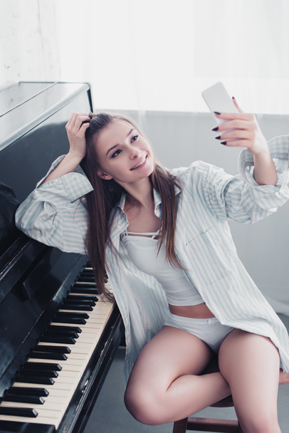 ελκυστικό κορίτσι στο πουκάμισο και εσώρουχα που κάθεται στο πιάνο και λαμβάνοντας αυτοπορτρέτα στο σαλόνι - Φωτογραφία, εικόνα