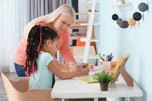 Female mature nanny helping little African-American girl to do homework - Photo, image