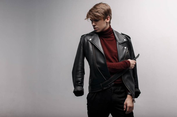 Handsome young attractive man with a hairstyle in a trendy black leather jacket in burgundy stylish golf and stylish black jeans is standing and posing in a room near a white wall. Modern guy on style - 写真・画像