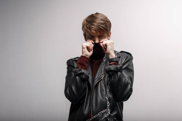 Молодой человек с модной прической в красном гольфе в кожаной куртке в черных джинсах закрывает лицо руками и смотрит в камеру в студии возле белой стены. Симпатичный парень.
. - Фото, изображение