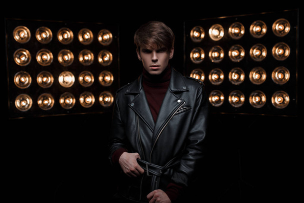 Attractive young stylish man in a leather jacket in a red stylish golf is standing and looking at the camera in a dark studio against the backdrop of bright orange electrical spotlights. Nice guy - Foto, imagen