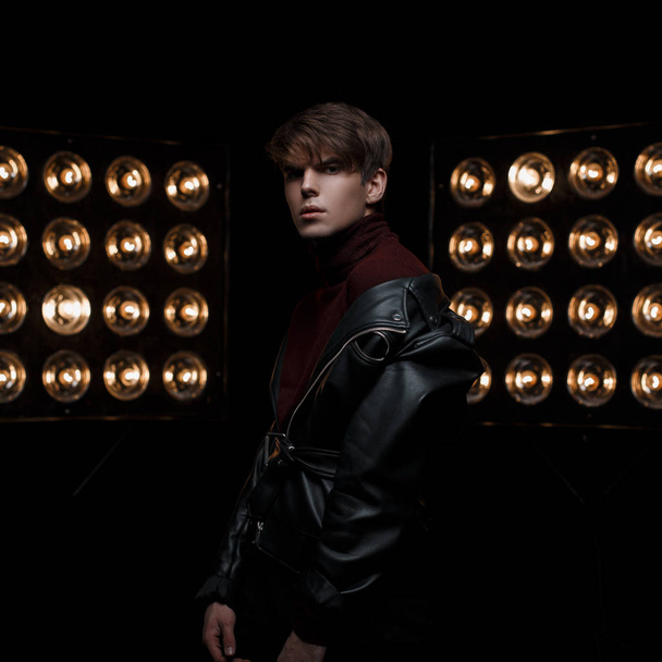 Serious mysterious young man in vintage trendy leather jacket in red stylish golf and black jeans is standing and posing in the studio near bright professional electric spotlights. Fashionable guy - Foto, Imagem