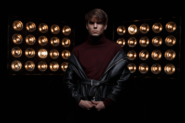 Attractive young stylish man in a leather jacket in burgundy stylish golf stands in a dark studio against the background of bright orange vintage paws of lights. Cute fashion star guy - 写真・画像