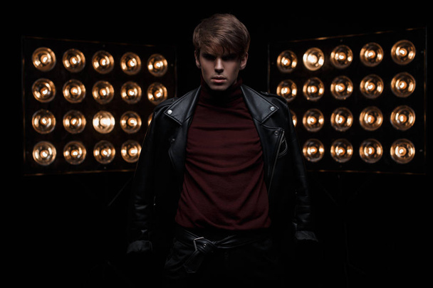 Young handsome fashionable man in a leather black jacket in burgundy golf in stylish pants posing in a dark studio against a background of bright electric vintage orange bulbs. Cute guy. Fashion. - Photo, Image