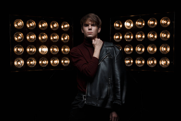 Pretty young handsome man in black stylish leather jacket in burgundy golf and black jeans poses looking into the camera in a dark room near bright vintage professional lamps. Stylish guy model - Foto, imagen