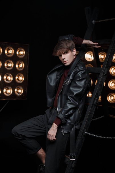 Young attractive man in a stylish leather jacket in retro style in jeans and golf posing in the studio near the metal stairs against the background of bright orange vintage lamps. Stylish sexy guy - 写真・画像