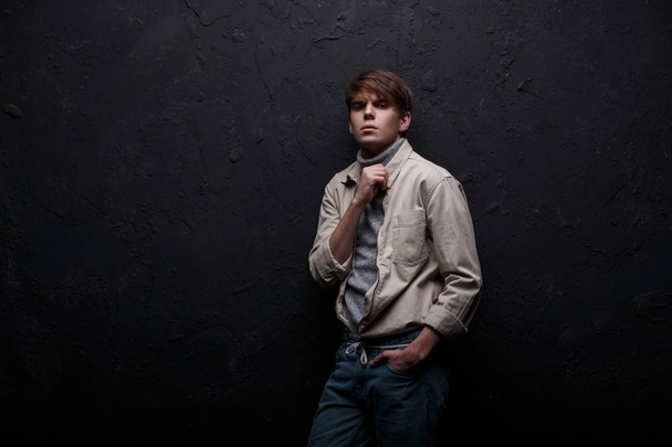 Stylish young man in a fashionable modern jacket in a gray sweater with a fashionable hairstyle in vintage jeans posing in a studio near a black wall. Handsome fashionable guy. American boy - Foto, Imagem