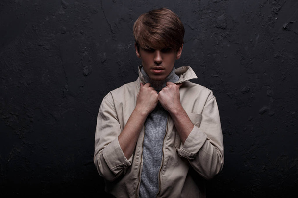 Young mysterious attractive man with a fashionable hairstyle in a spring light jacket in a vintage sweater standing and looking at the camera in a room near the black wall. Fashionable handsome guy - Foto, afbeelding