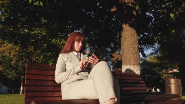 womanbusinessman wearing glasses and light suit works with tablet and checks email in summer park on bench lit by the bright evening sun - Video, Çekim