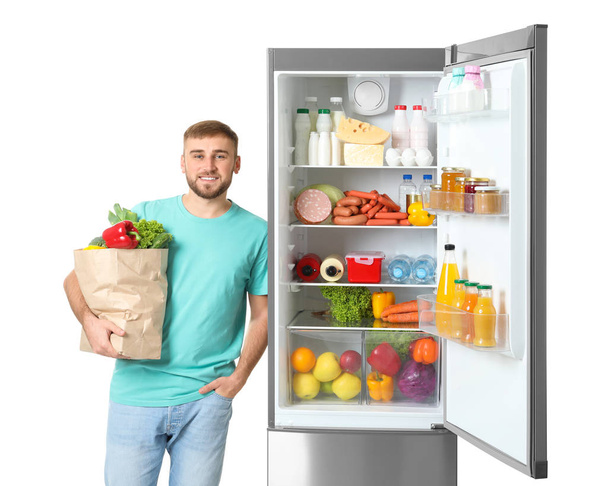 Young man with bag of groceries near open refrigerator on white background - Фото, изображение