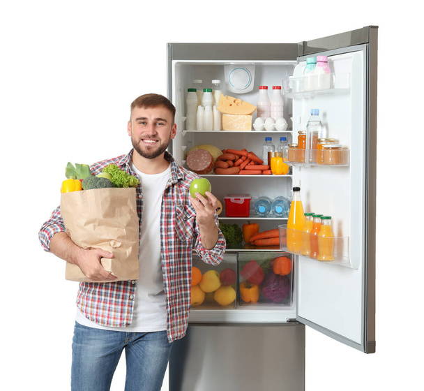 Young man with bag of groceries and apple near open refrigerator on white background - Foto, Imagem