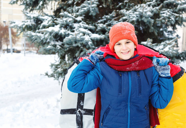 赤い帽子スカーフ、青ジャケットの陽気なかわいい少年雪のチューブを保持は、楽しく、笑顔です。冬の公園でソリのティーネー ジャー。アクティブなライフ スタイル、冬の活動、屋外の冬のゲーム - 写真・画像