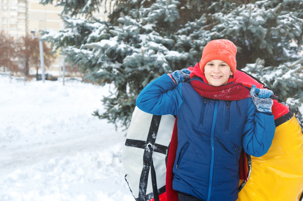веселый милый мальчик в шляпе красный шарф и синяя куртка держит трубку на снегу, веселится, улыбается. Подросток на санях в зимнем парке. активный образ жизни, зимняя активность, открытый зимний игры
 - Фото, изображение