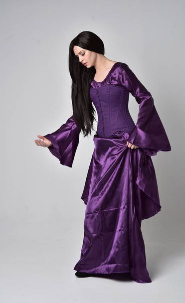 portrait pleine longueur de belle fille aux longs cheveux noirs, portant une robe médiévale fantaisie violette. pose debout sur fond de studio gris
. - Photo, image