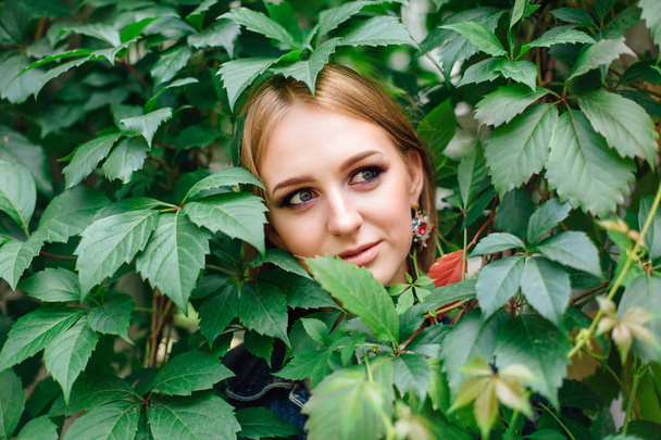 Portrait of a young beautiful woman with make up and ear rings, hiding in leaves of grapes. - Fotoğraf, Görsel