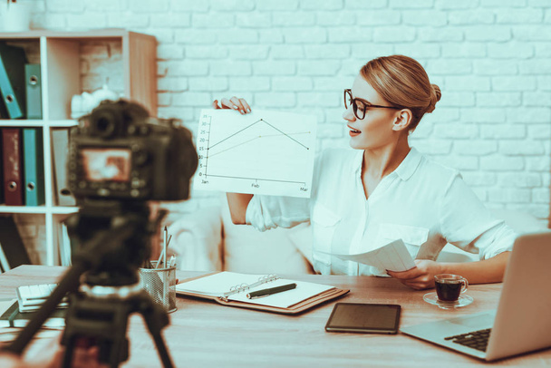 Blogger Makes a Video. Blogger is Businesswoman. Video About a Business. Operator Shoots a Video on Camera. Laptop and Supplies on Table. Woman Showing a Business Graph. People in Studio Interior. - 写真・画像