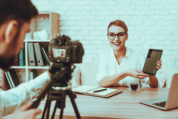 Blogger Makes a Video. Blogger is Smiling Businesswoman. Video About a Business. Man Operator Shoots a Video on Camera. Laptop and Supplies on Table. Woman Showing a Tablet. People in Studio Interior. - Zdjęcie, obraz