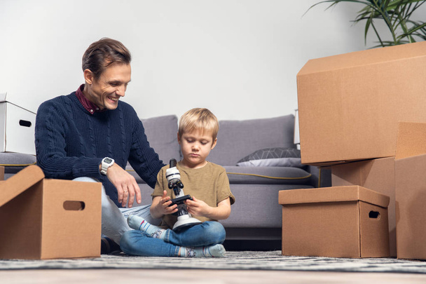 Photo of man and small boy with microscope sitting on floor among cardboard boxes - Φωτογραφία, εικόνα