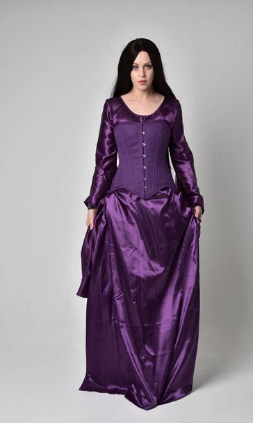 retrato de longitud completa de hermosa chica con el pelo largo negro, usando vestido medieval de fantasía púrpura. pose de pie sobre fondo gris estudio
. - Foto, imagen