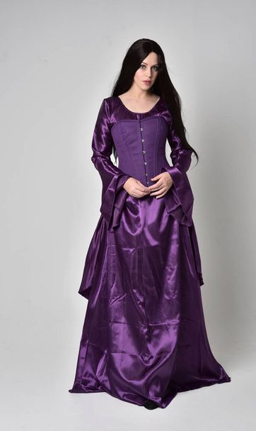 Ganzkörperporträt eines schönen Mädchens mit langen schwarzen Haaren, das ein lila mittelalterliches Fantasiegewand trägt. Stehende Pose auf grauem Studiohintergrund. - Foto, Bild