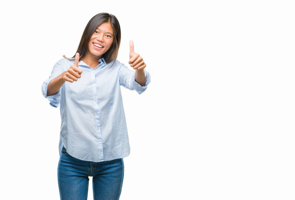 Young asian business woman over isolated background approving doing positive gesture with hand, thumbs up smiling and happy for success. Looking at the camera, winner gesture. - Photo, image