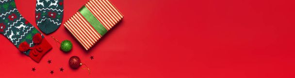 Шерстяные носки с новогодним орнаментом, подарочная коробка, шарики, блестки конфетти на красном фоне сверху вид плоский лежал. Концепция праздника, представляет Рождество. Поздравления фон с пространством для текста
 - Фото, изображение