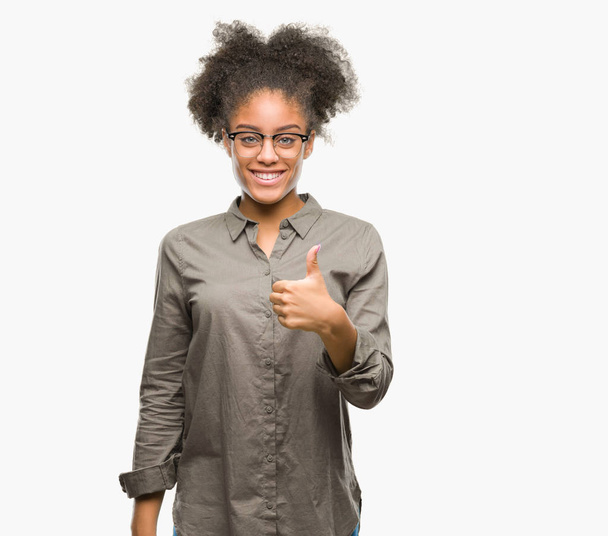 Mujer afroamericana joven con gafas sobre un fondo aislado haciendo un gesto de pulgares felices hacia arriba con la mano. Aprobar la expresión mirando a la cámara con éxito
. - Foto, imagen