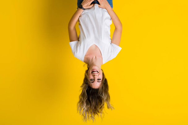 Beautiful young blonde woman jumping happy and excited hanging upside down over isolated yellow background - Photo, Image