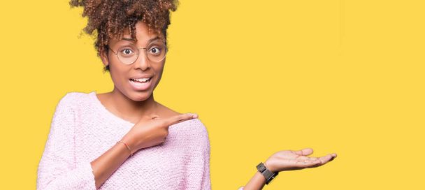 Красива молода афро-американських жінка в окулярах над ізольованих фон здивували і посміхається на камеру під час представлення рукою і вказуючи пальцем. - Фото, зображення