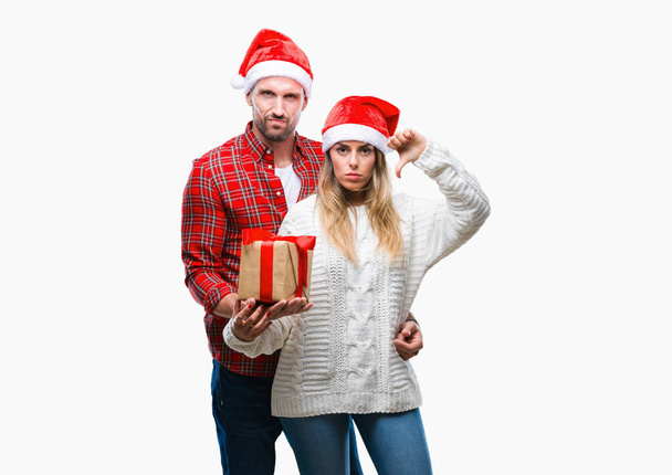 Jeune couple amoureux portant le chapeau de Noël et tenant présent sur fond isolé avec le visage en colère, signe négatif montrant aversion avec les pouces vers le bas, concept de rejet
 - Photo, image