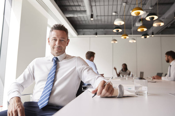 Portrait Of Mature Businessman Sitting In Modern Boardroom With Colleagues Meeting Around Table In Background - Foto, Bild