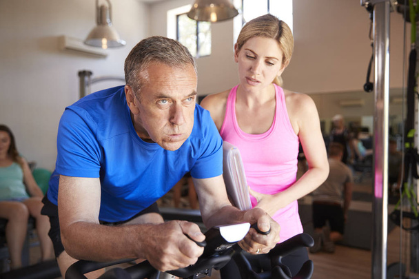 Senior Man Exercising On Cycling Machine Being Encouraged By Female Personal Trainer In Gym - Fotoğraf, Görsel