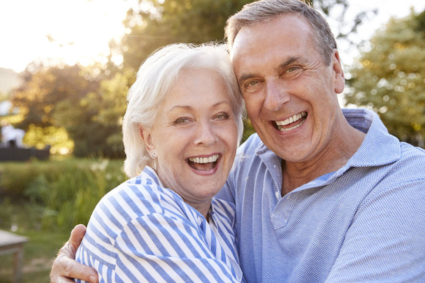 Portrait Of Loving Senior Couple Hugging Outdoors In Summer Park Against Flaring Sun - Foto, imagen