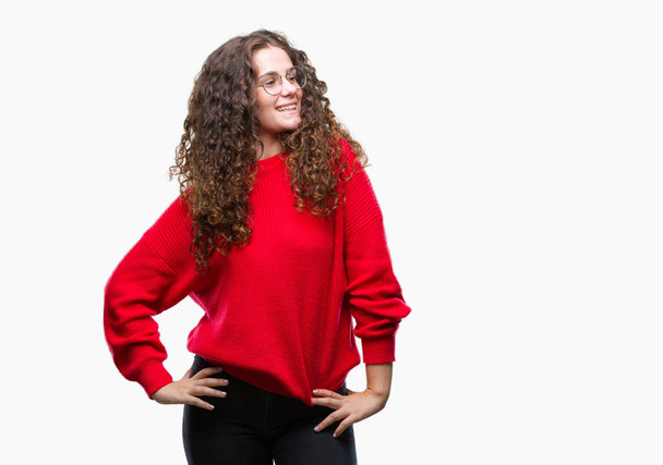 Beautiful brunette curly hair young girl wearing glasses and winter sweater over isolated background looking away to side with smile on face, natural expression. Laughing confident. - Photo, image