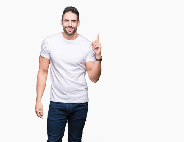 Hombre joven con una camiseta blanca casual sobre un fondo aislado con una gran sonrisa en la cara, apuntando con la mano y el dedo hacia el lado mirando a la cámara
. - Foto, Imagen