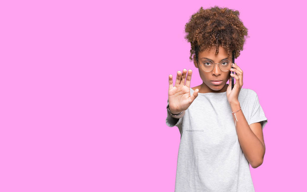 Mujer afroamericana joven hablando en smartphone sobre fondo aislado con la mano abierta haciendo stop sign con expresión seria y segura, gesto de defensa
 - Foto, imagen