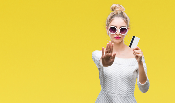 Mladá krásná blondýna žena držící kreditní karty nad izolované pozadí s otevřenou rukou dělá stopku s vážnou a sebevědomý výraz, gesto obrany - Fotografie, Obrázek